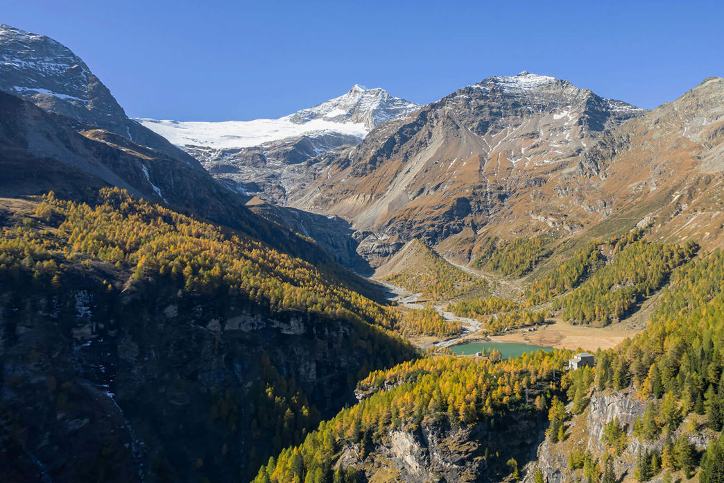 Panorama alpino con ghiacciaio, bosco di larici e piccolo lago in una valle di montagna autunnale.