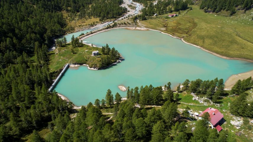 Luftaufnahme des türkisfarbenen Palüsees mit Staumauer, eingebettet in eine bewaldete Berglandschaft.