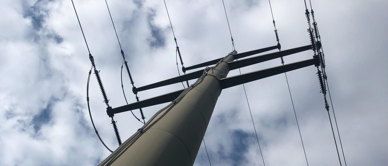 Blick von unten auf einen Strommast mit Hochspannungsleitungen vor bewölktem Himmel.
