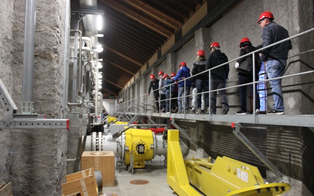Besuchergruppe mit Helmen bei Kraftwerksführung auf Galerie oberhalb technischer Anlagen.