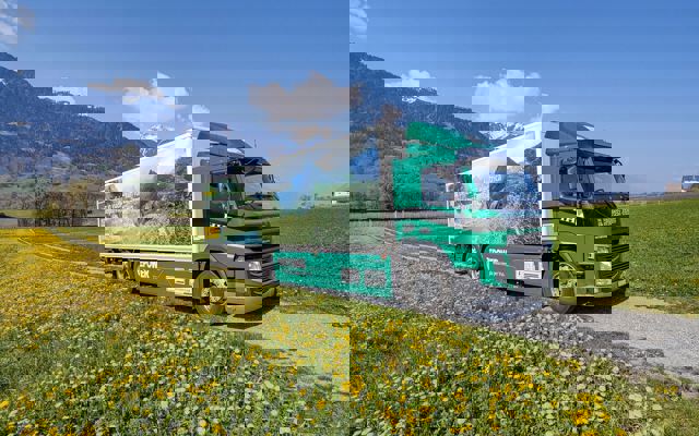 Elektro-LKW von Hunger fährt durch eine blühende Wiese vor alpiner Landschaft und blauem Himmel.